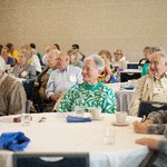 Multiple people are seated at tables and paying attention to a point not shown in the picture